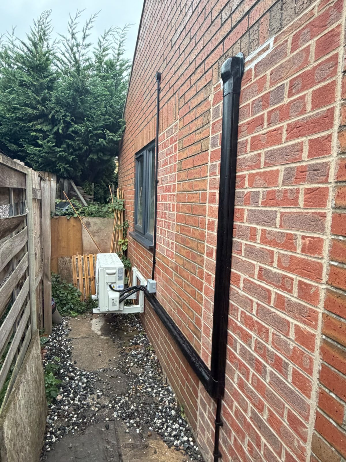 Outdoor air conditioning unit on a wall bracket at the side of the brick garden room, with black trunking along the wall