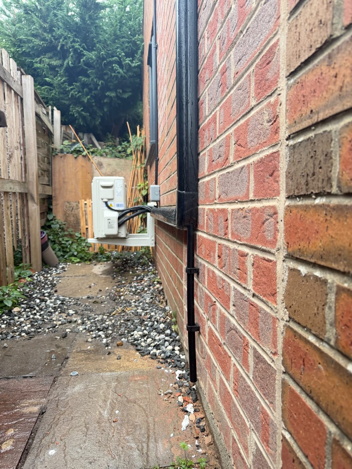Narrow side passage with the wall-mounted outdoor unit and black trunking running down the brickwork